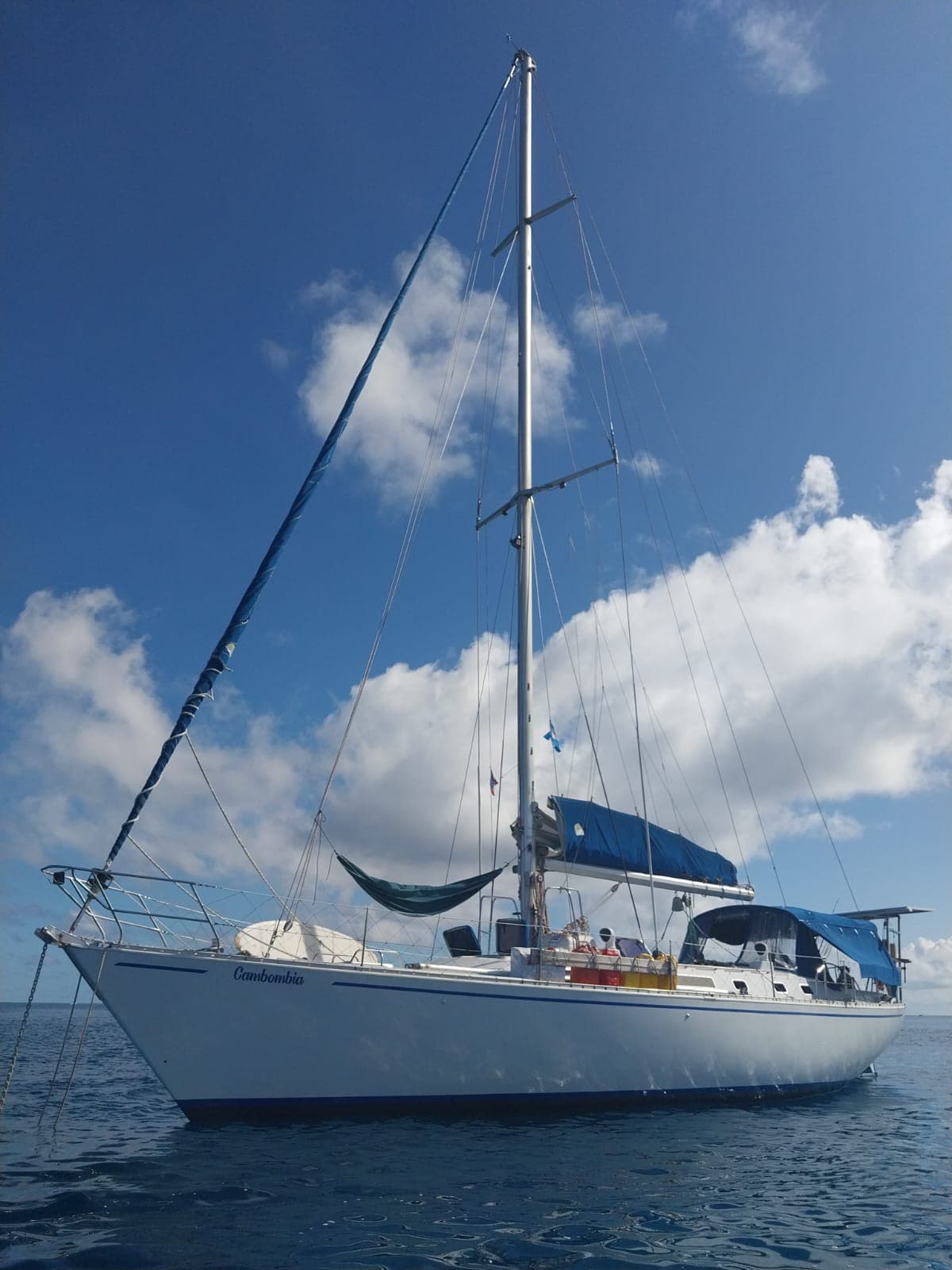 A sailing vessel is anchored on calm blue waters under a partly cloudy sky. The boat features a tall mast with sails furled and a canopy providing shade. A small group of people can be seen enjoying the deck, creating a relaxed atmosphere.
