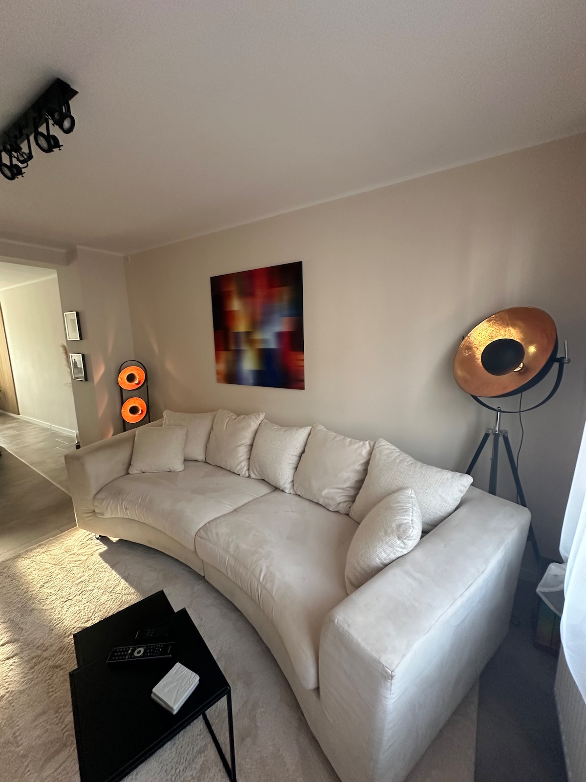 A spacious curved white couch is positioned in a well-lit living area. A colorful abstract artwork adds visual interest to the wall. Modern lighting fixtures, including a stylish floor lamp, enhance the room's contemporary design, creating a comfortable atmosphere.
