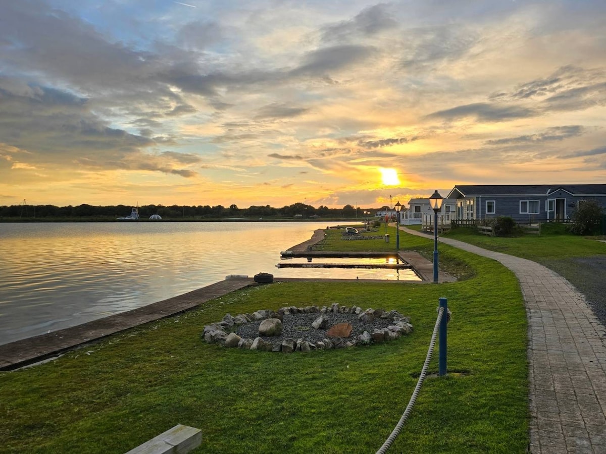 A serene lakeside view captures a colorful sunset over calm waters, with a stone fire pit surrounded by grass in the foreground. A pathway lined with grass and a dock are visible alongside the water, leading to charming lodges in the distance.