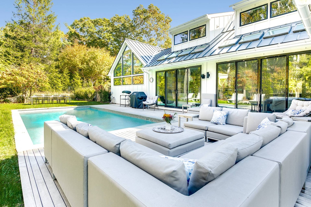 An outdoor seating area is arranged around a central table, featuring a spacious sectional sofa with soft cushions. A heated saltwater pool glistens nearby, surrounded by a deck and lush greenery, with large glass doors leading into the modern farmhouse.