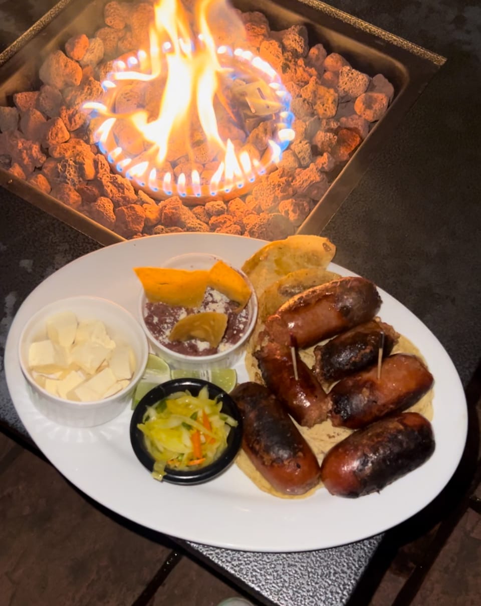 A plate of grilled sausages is arranged with accompaniments, including cheese cubes, pickled vegetables, and a small serving of sauce. A flame flickers in the background, providing a warm glow, while a dark surface outlines the meal.
