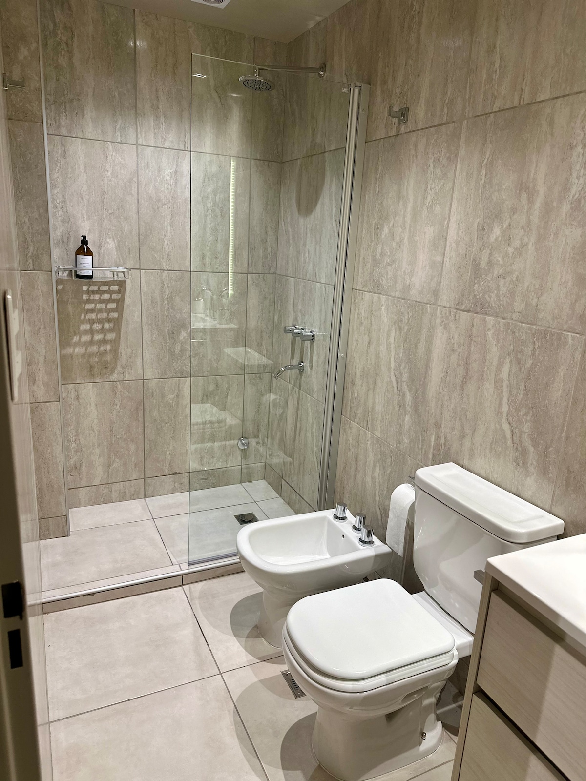 A modern bathroom is depicted, featuring a glass-enclosed shower with a rain showerhead. Light-colored tiles adorn the walls and floor. A white toilet is positioned beside a bidet, with a sleek vanity unit visible in the frame.