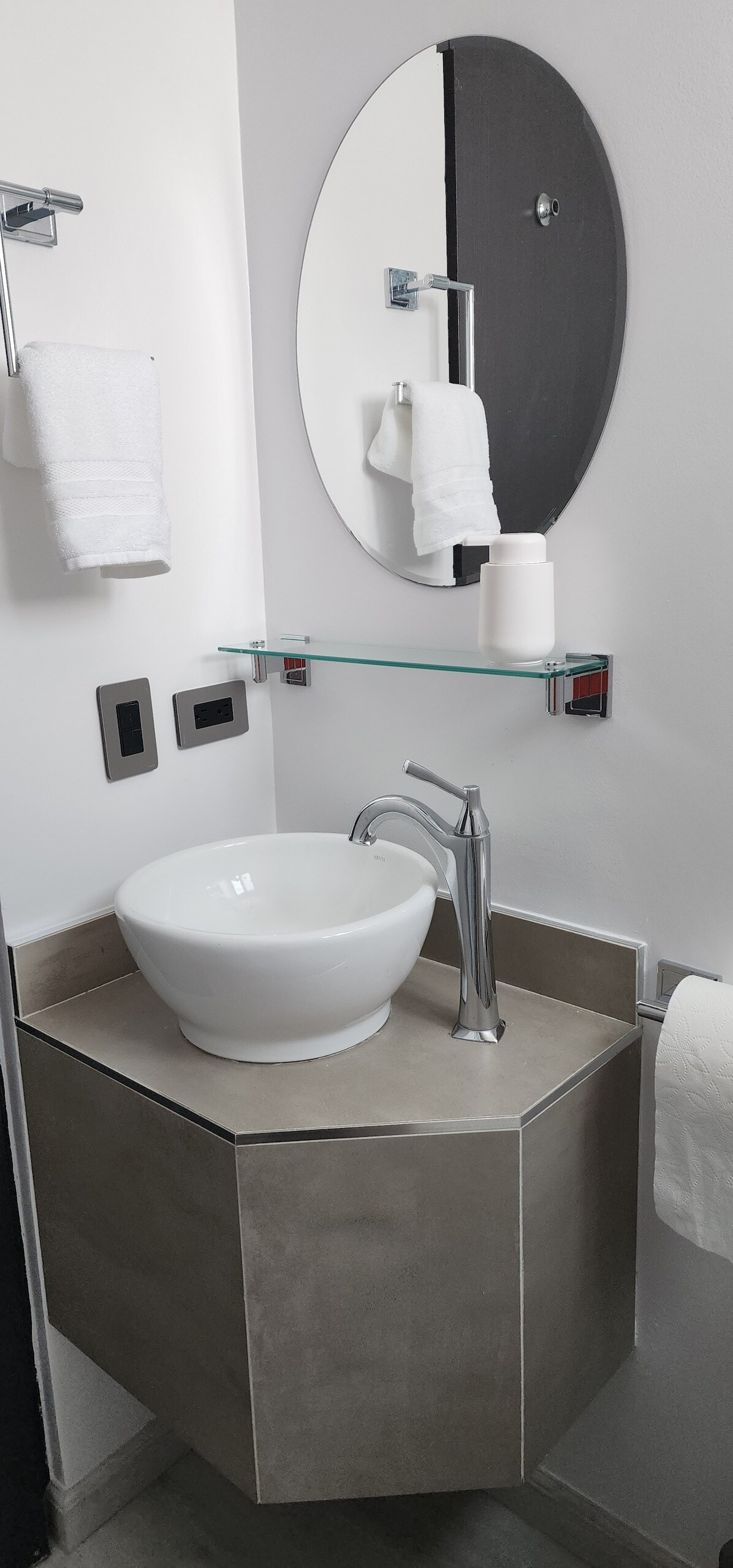 A compact bathroom is presented, featuring a contemporary basin with a sleek faucet. A circular mirror is mounted above the sink, complemented by glass shelving and neatly arranged towels. Minimalist fixtures enhance the overall modern design.
