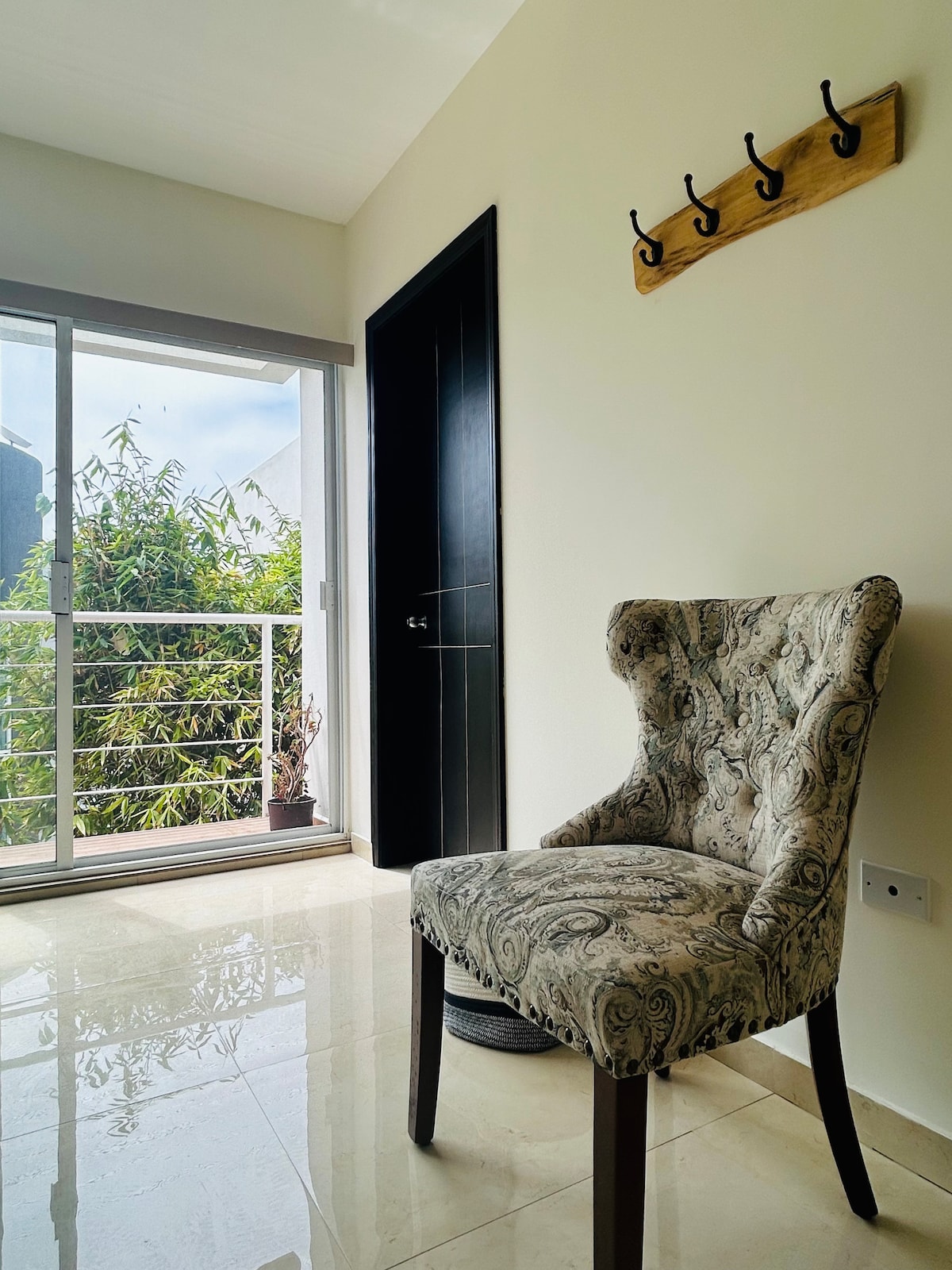 A well-lit entryway features a patterned upholstered chair positioned beside a sliding glass door that leads to a small balcony. Natural light fills the space, highlighting the polished floor and a wall-mounted hook rack, while greenery is visible through the glass.