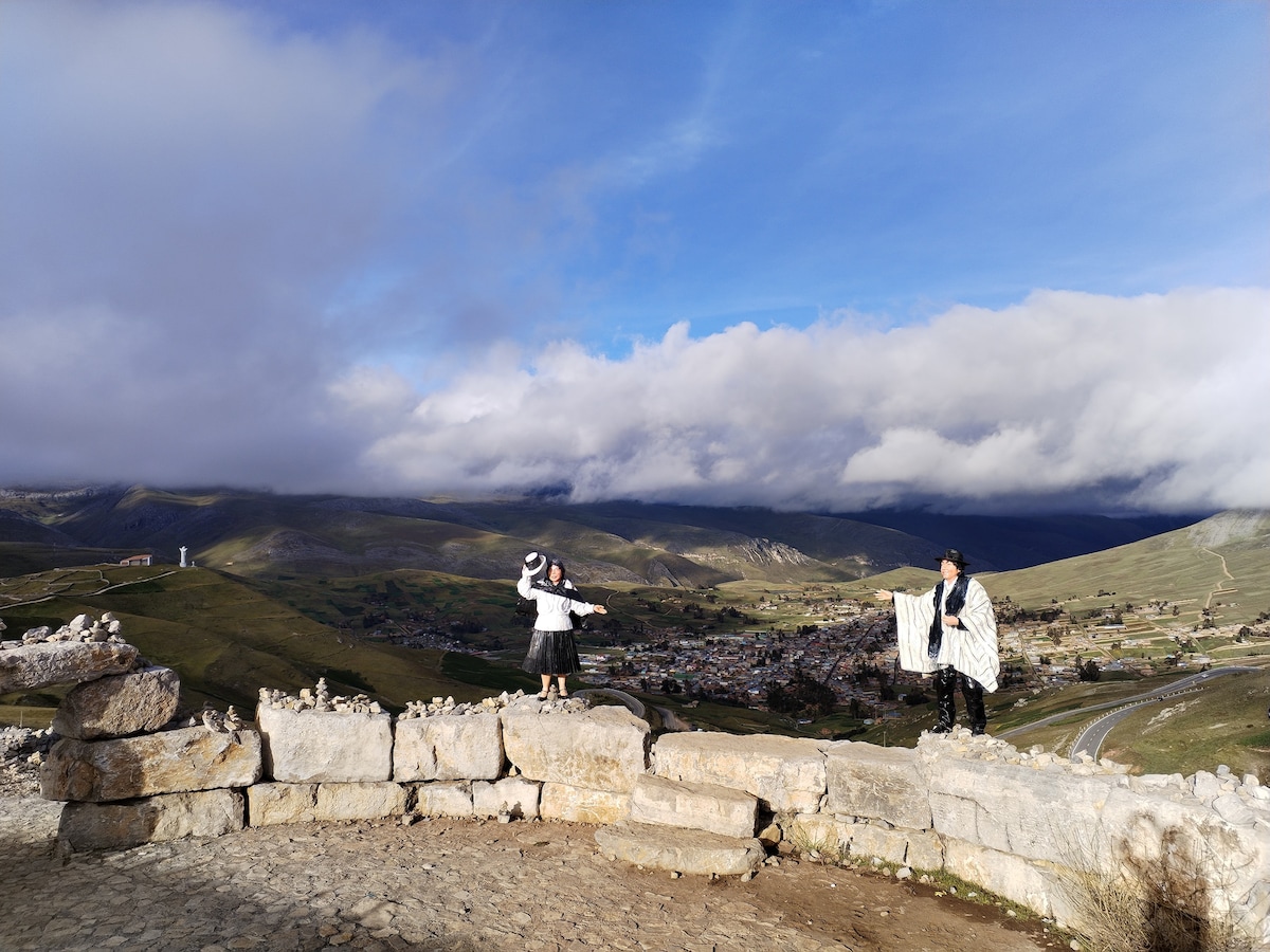 A scenic landscape is depicted, with two individuals standing on a stone wall. Lush green hills and a small town are visible in the background under a partly cloudy sky. The scene captures a blend of nature and culture, showcasing the beauty of the surrounding area.