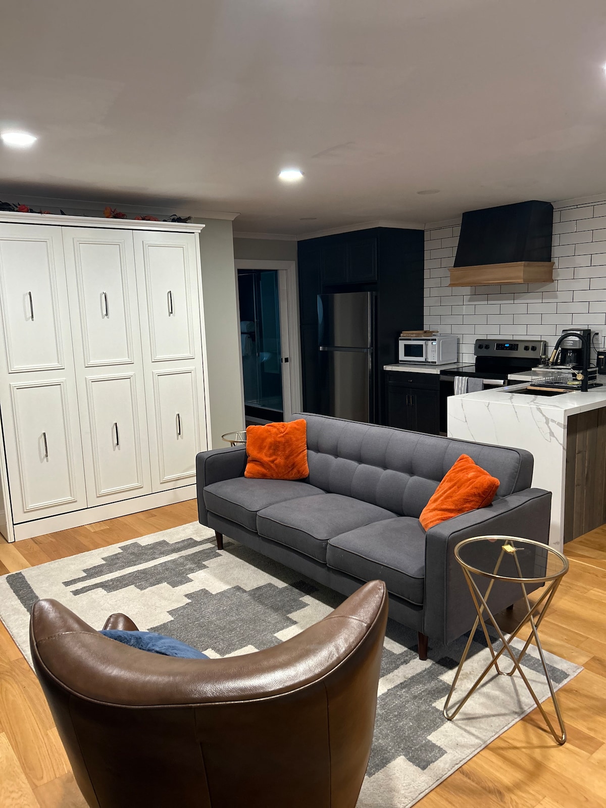 A modern living space features a gray sofa adorned with orange throw pillows, paired with a brown accent chair. A light area rug with geometric patterns is situated on warm wooden flooring. A kitchen area, seen in the background, includes dark cabinetry and stainless-steel appliances.