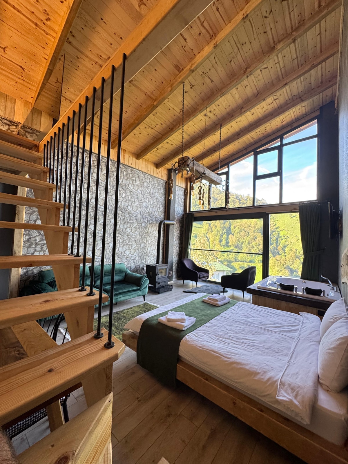 A spacious room features high wooden ceilings and large windows that invite natural light and views of the surrounding greenery. A comfortable bed is complemented by a cozy seating area with two chairs and a sofa. A wooden staircase leads to an upper level.