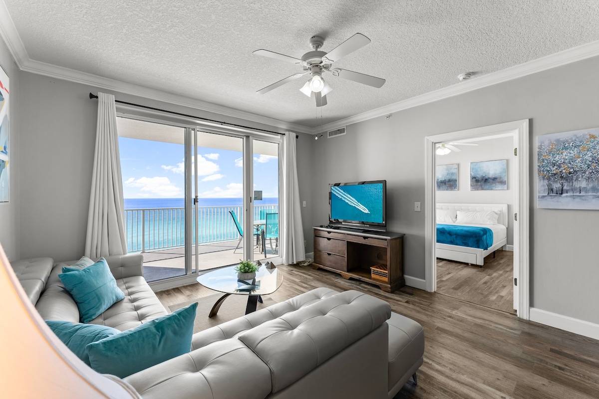 A spacious living area features a light-colored sectional sofa with decorative cushions. A flat screen TV is situated on a modern console. Large glass doors open to a private balcony, revealing expansive views of the ocean and sky. Natural light illuminates the room.