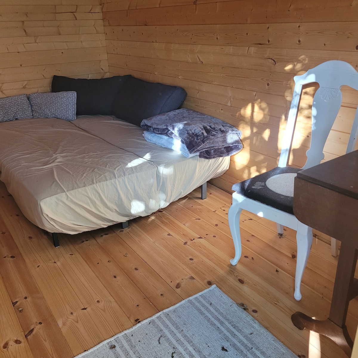 An inviting sleeping area features a spacious bed adorned with soft blankets and cushions. A simple wooden chair and a small writing desk complete the space. Natural light filters in, illuminating the wooden walls and flooring, enhancing the cozy ambiance.