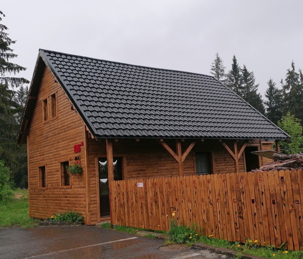 A rustic wooden house features a sloped roof and multiple windows, surrounded by a wooden fence. Its natural exterior is enhanced by lush greenery in the background, indicative of a tranquil setting. The entrance is visible, offering a welcoming facade.