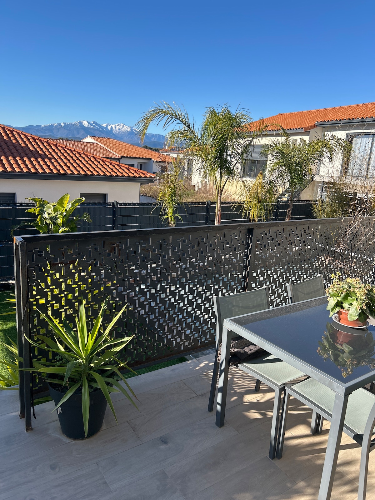 A sunlit terrace features a glass-top dining table surrounded by four chairs. Lush potted plants are present, adding greenery to the space. In the background, residences are visible along with distant mountains under a clear blue sky.