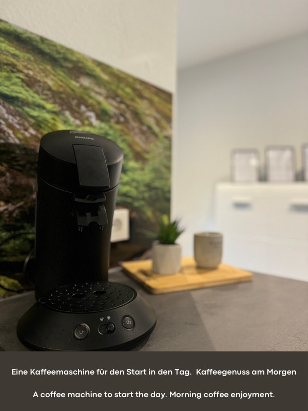 A coffee machine is positioned on a countertop, accompanied by a wooden tray featuring small items for coffee preparation. The surrounding area is designed with neutral tones and natural elements, suggesting a functional space for morning routines.