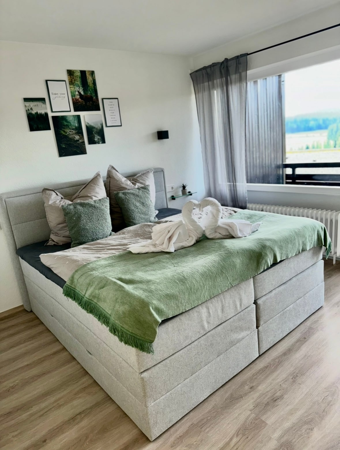 A spacious bed is dressed with soft, earthy linens, including decorative pillows and a light green throw. Natural light filters through sheer curtains, casting a gentle glow on the comfort of the room. Scenic views of the surrounding landscape are visible through the balcony window.