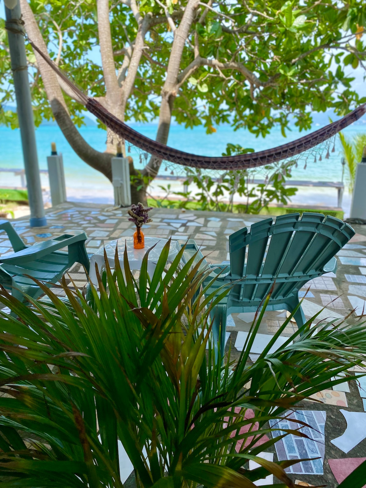 A serene outdoor space features two blue adirondack chairs positioned on a tiled patio, surrounded by lush greenery. A hammock hangs between two trees, overlooking the calm blue waters, creating an inviting area for relaxation near the beach.