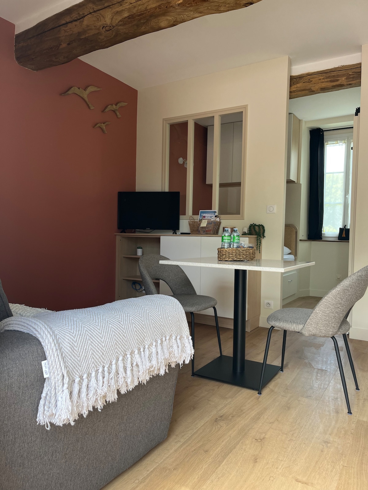 An inviting living area features a soft couch draped with a textured blanket. A small dining table with two chairs is positioned near a window, allowing natural light to fill the space. The wall displays subtle decor and wooden beams add character to the ceiling.