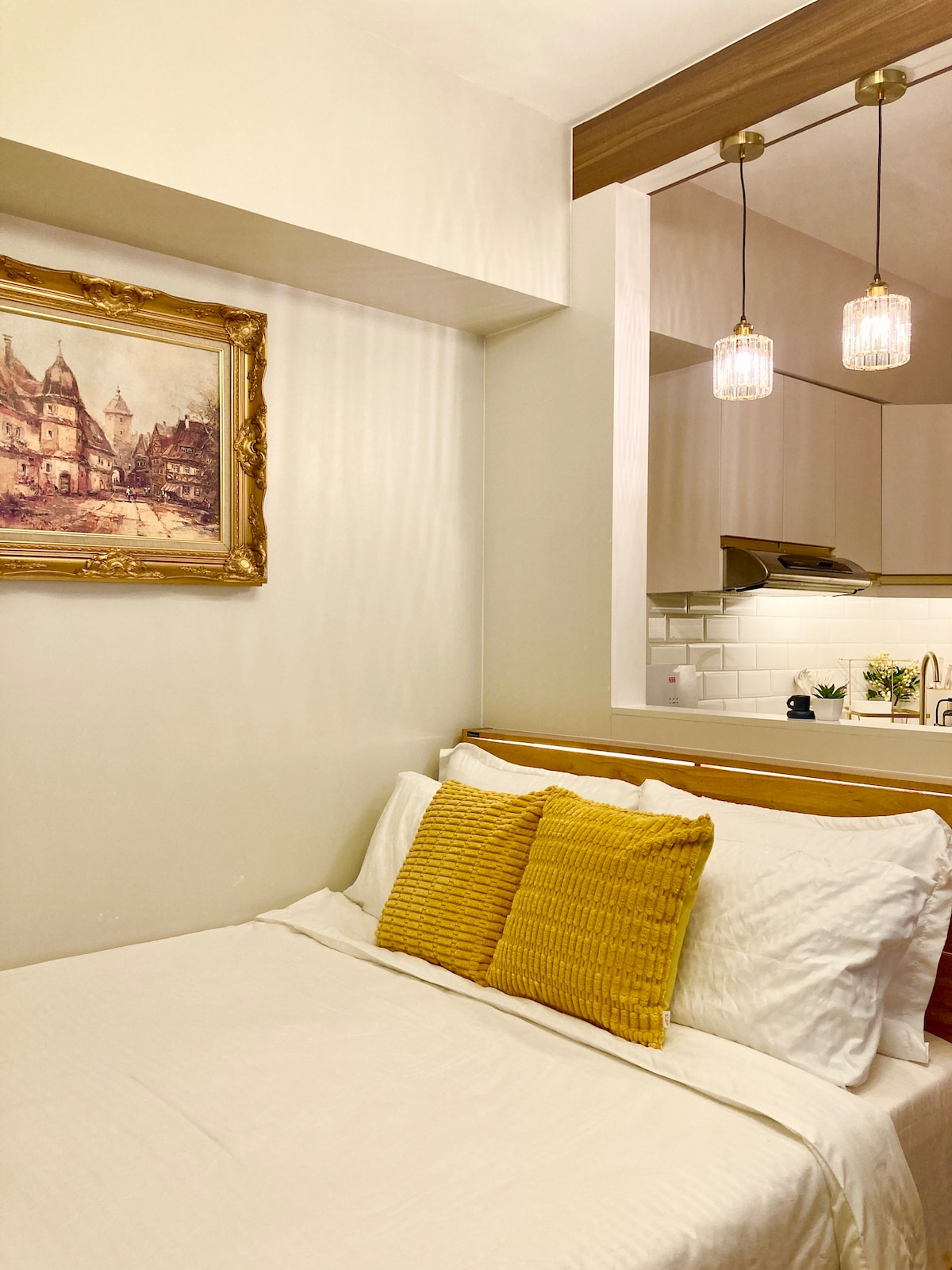 A cozy sleeping area is highlighted by a queen-sized bed adorned with white linens and two yellow decorative pillows. A vintage painting in a gold frame hangs on the wall, complemented by warm pendant lighting above the kitchenette.