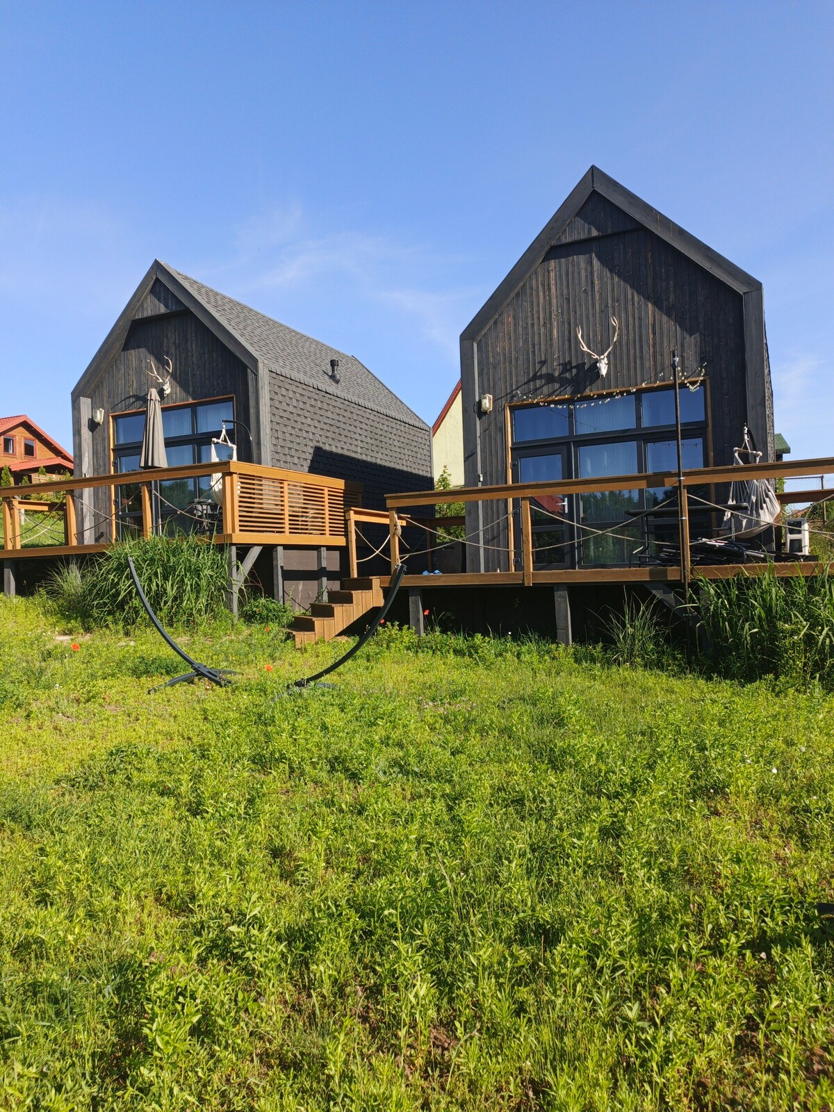 The image shows two modern cottages with contrasting dark and light exteriors. They feature large windows and patios with wooden railings, surrounded by lush green grass. The design reflects a contemporary barn style, emphasizing a connection with nature.
