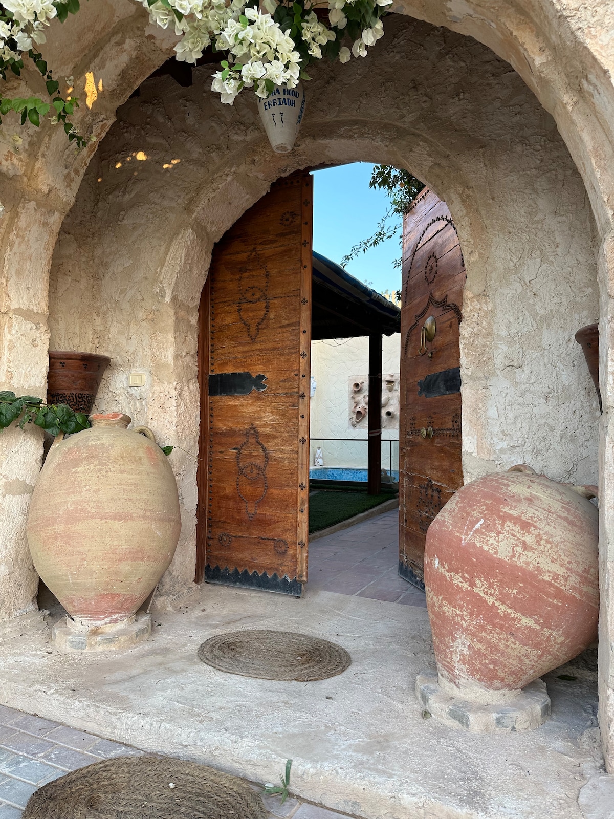 An arched entrance features wooden doors adorned with intricate carvings. Flanking the doorway are two large, decorative clay pots. Natural light filters through, illuminating the textured wall and the inviting passage leading into the home.