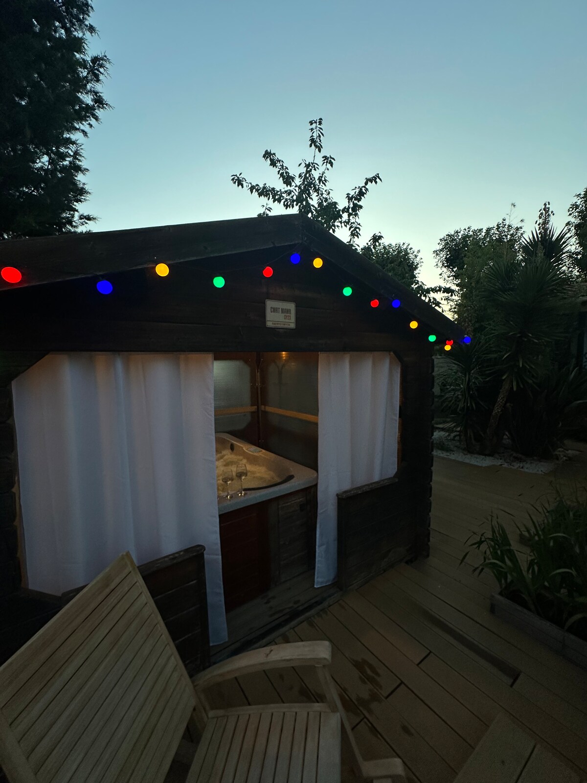 A cozy wooden structure housing a jacuzzi is adorned with colorful string lights. Soft white curtains provide privacy, while the surrounding greenery adds a touch of nature. A wooden seating area is positioned nearby on the deck, inviting relaxation.