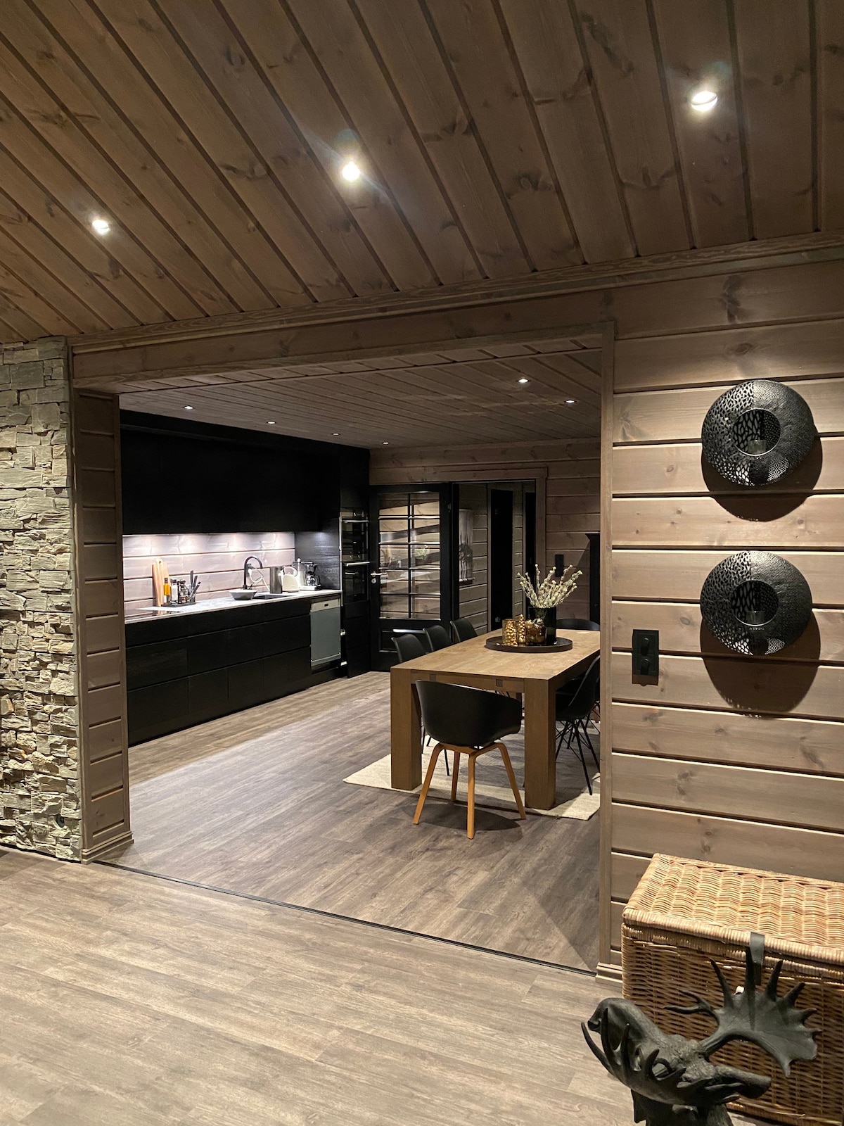 A modern kitchen and dining area are visible through an archway, featuring a long wooden dining table surrounded by black chairs. Sleek cabinetry in dark tones is complemented by a contrasting light backsplash, while stylish decorative items add visual interest.