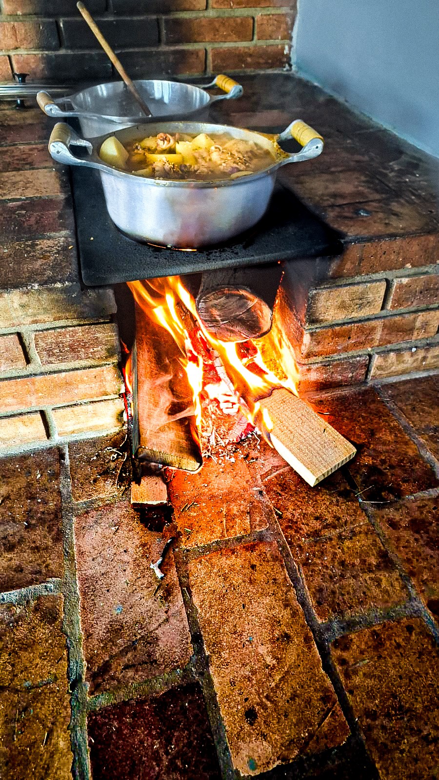 A rustic area is shown with a chimney stacked with bricks, where a fire burns beneath a metal stove. Two pots of food sit above the flames, with smoke rising gently, indicating a warm cooking environment.