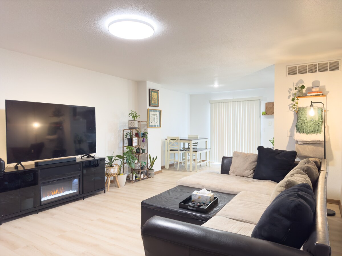 The living room showcases a large sectional sofa in a neutral color, accented by a modern coffee table. A sizable TV is mounted on the wall, with a media console beneath. Soft lighting enhances the open space, leading to a dining area in the background.