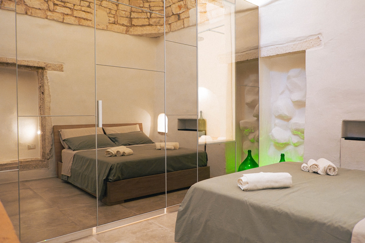 The bedroom features a spacious bed with neutral bedding, framed by a striking mirrored wall. Soft lighting creates a calm atmosphere, while a decorative alcove with textured stone adds character. Complementary green glass decor pieces enhance the room's unique design.