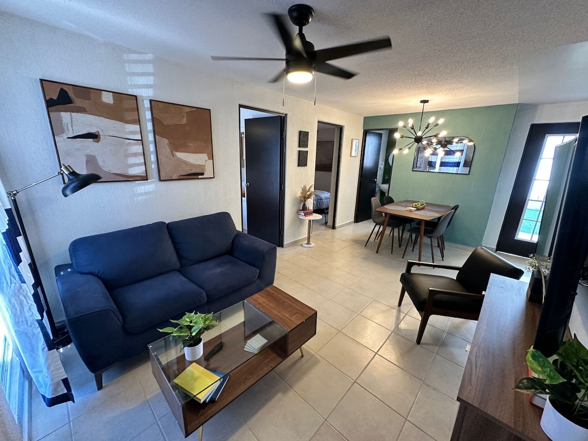 A modern living area features a navy blue sofa, a wooden coffee table, and a dark wood entertainment unit. A ceiling fan is mounted above, while the dining area showcases a round table with four chairs. Large windows provide natural light.