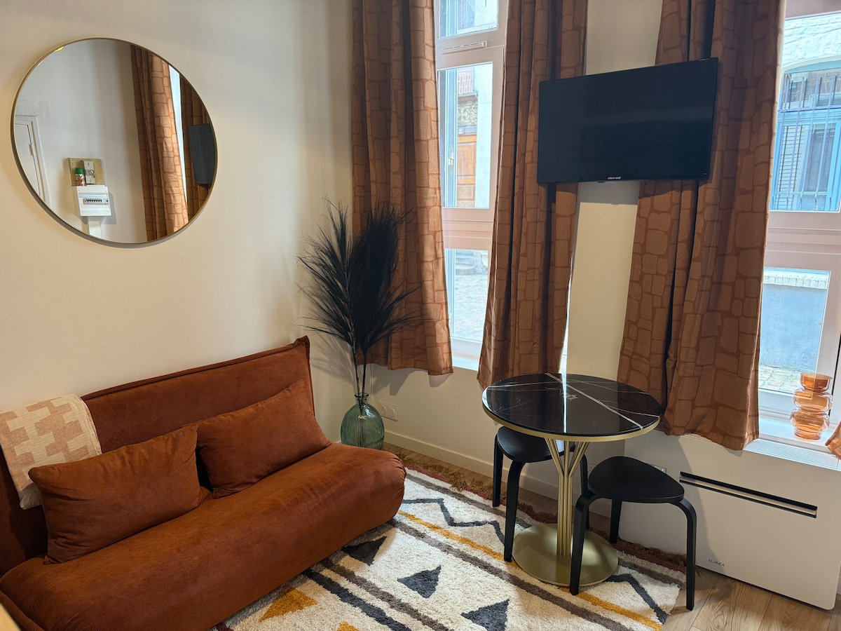 A cozy seating area is highlighted by a brown sofa and a circular mirror above it. A small round dining table is positioned near the window, accompanied by a black television mounted on the wall. Tall curtains frame the window, and a textured area rug adds warmth to the space.