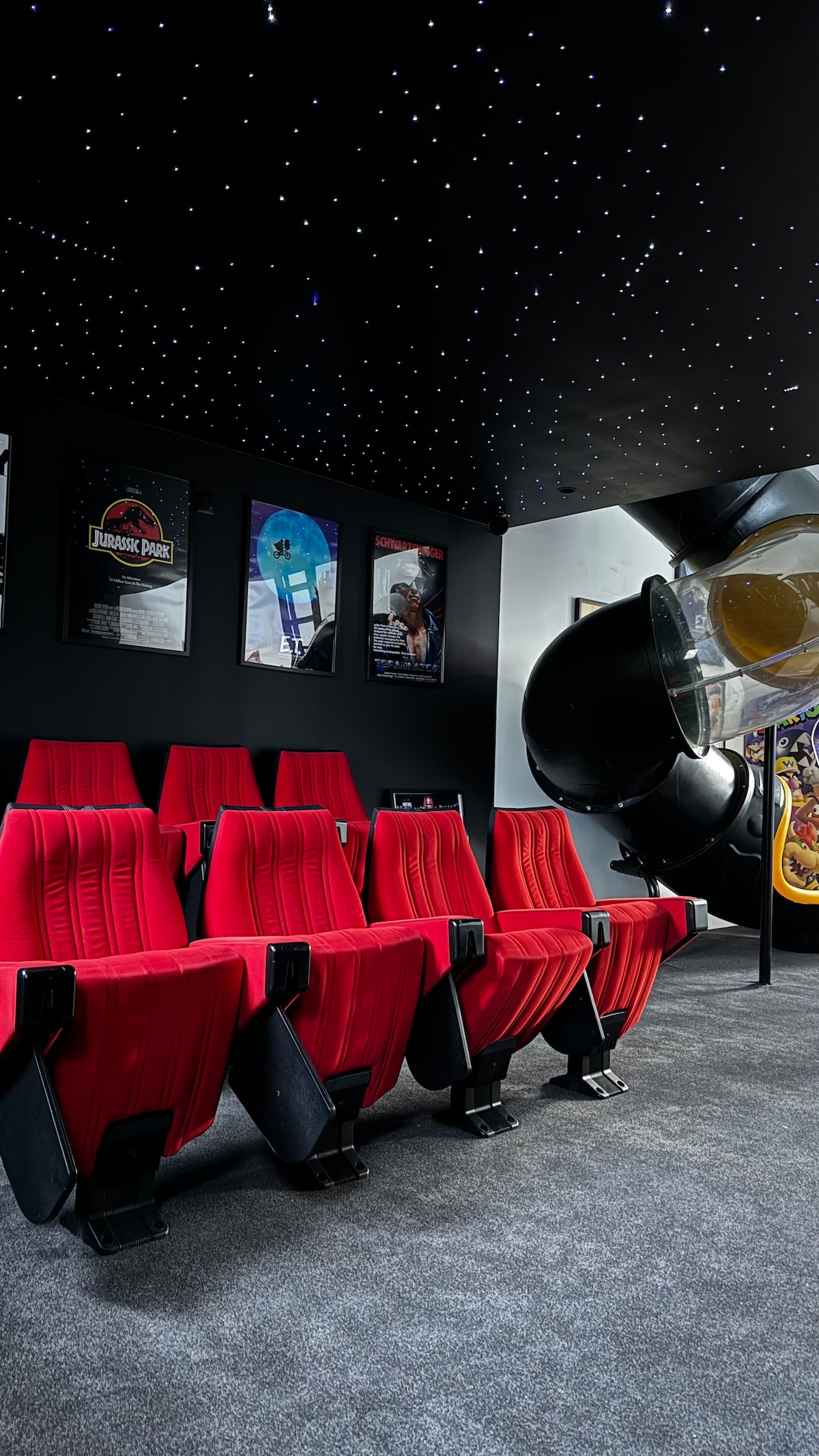 A private cinema room features red theater seats arranged in a viewing area, complemented by a starlit ceiling. Movie posters adorn the walls, creating an immersive entertainment environment. The space is designed for comfortable movie nights with friends and family.