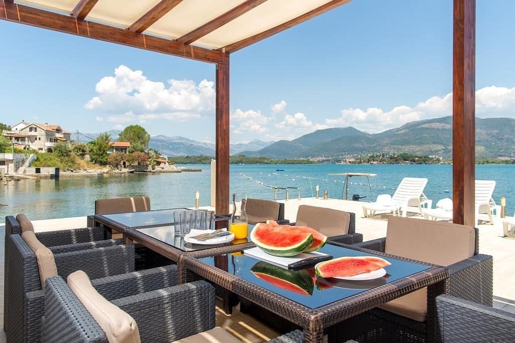 An outdoor dining area is presented under a shaded pergola, featuring a large table with comfortable seating. Fresh watermelon slices and drinks are displayed on the table. The scenic backdrop includes a calm bay and distant mountains, providing a serene atmosphere.