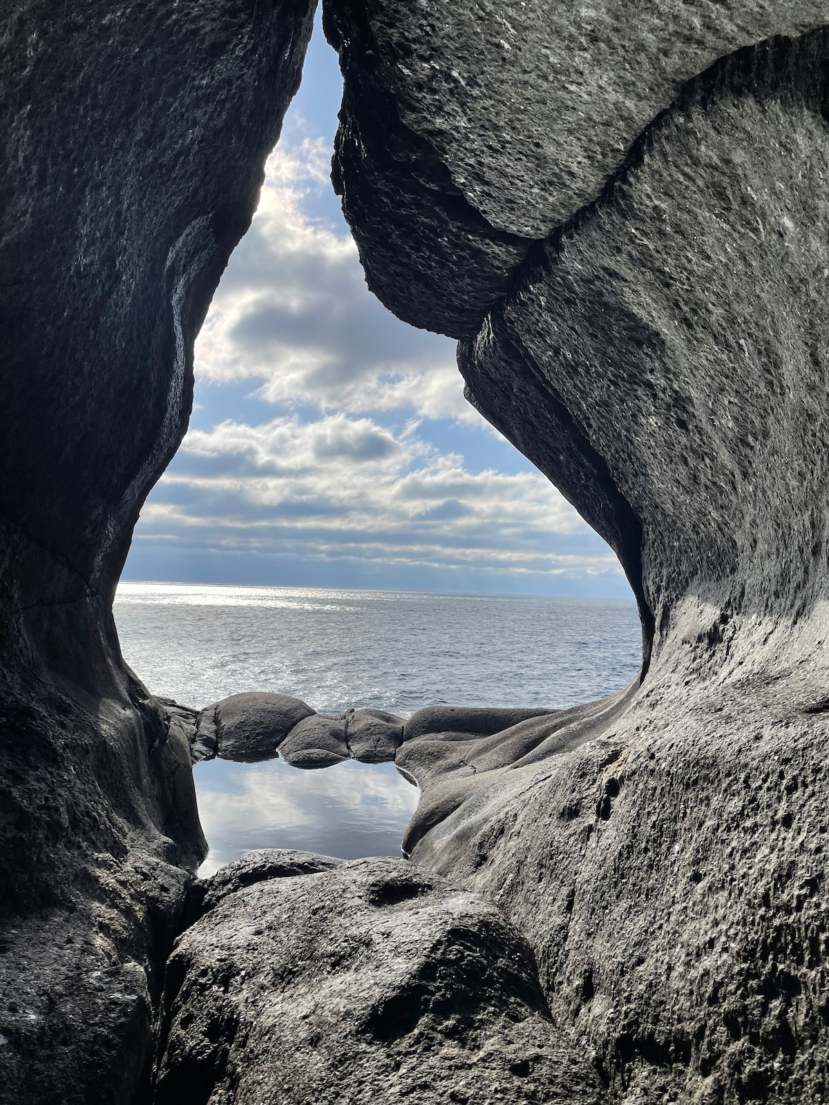 A natural rock formation frames a view of the calm sea and an overcast sky. The water reflects light, creating a serene atmosphere, while the rugged texture of the rocks adds depth to the scene.
