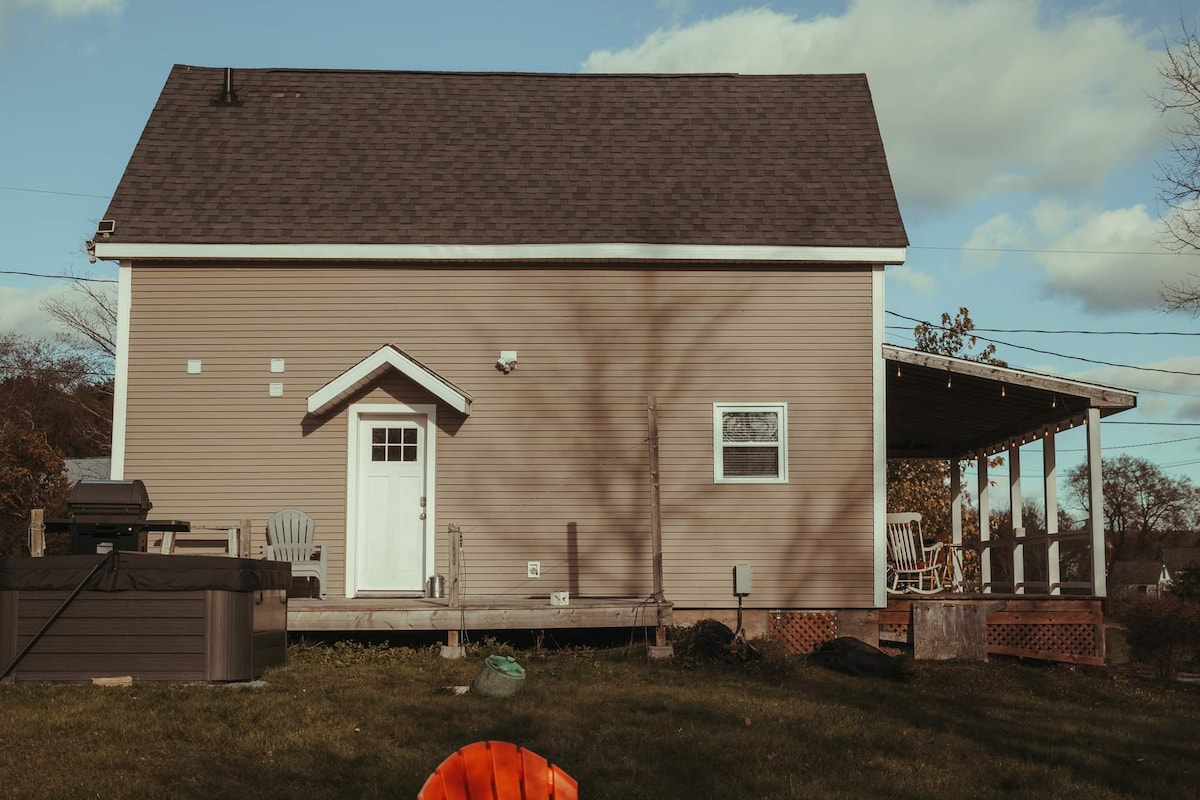 The exterior of a two-story house is presented, showcasing a light brown facade under a blue sky. A wooden deck with seating is located to the right, featuring a porch roof that provides shade. A storage area is visible to the left, with outdoor furniture arranged for relaxation.