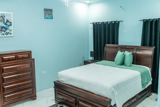 A serene sleeping area features calming blue walls accented by a piece of floral wall art. A spacious bed with muted green pillows is complemented by dark curtains and wooden furniture, including a dresser and nightstand. Soft lighting enhances the room's tranquil ambiance.