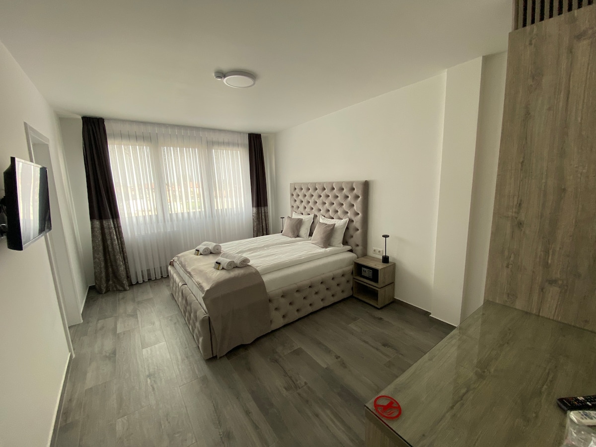 A modern bedroom is presented with a spacious bed featuring a tufted headboard. Natural light filters through sheer curtains, illuminating the neutral-toned decor. A stylish bedside table and a flat-screen television are positioned for convenience, enhancing the relaxing environment.
