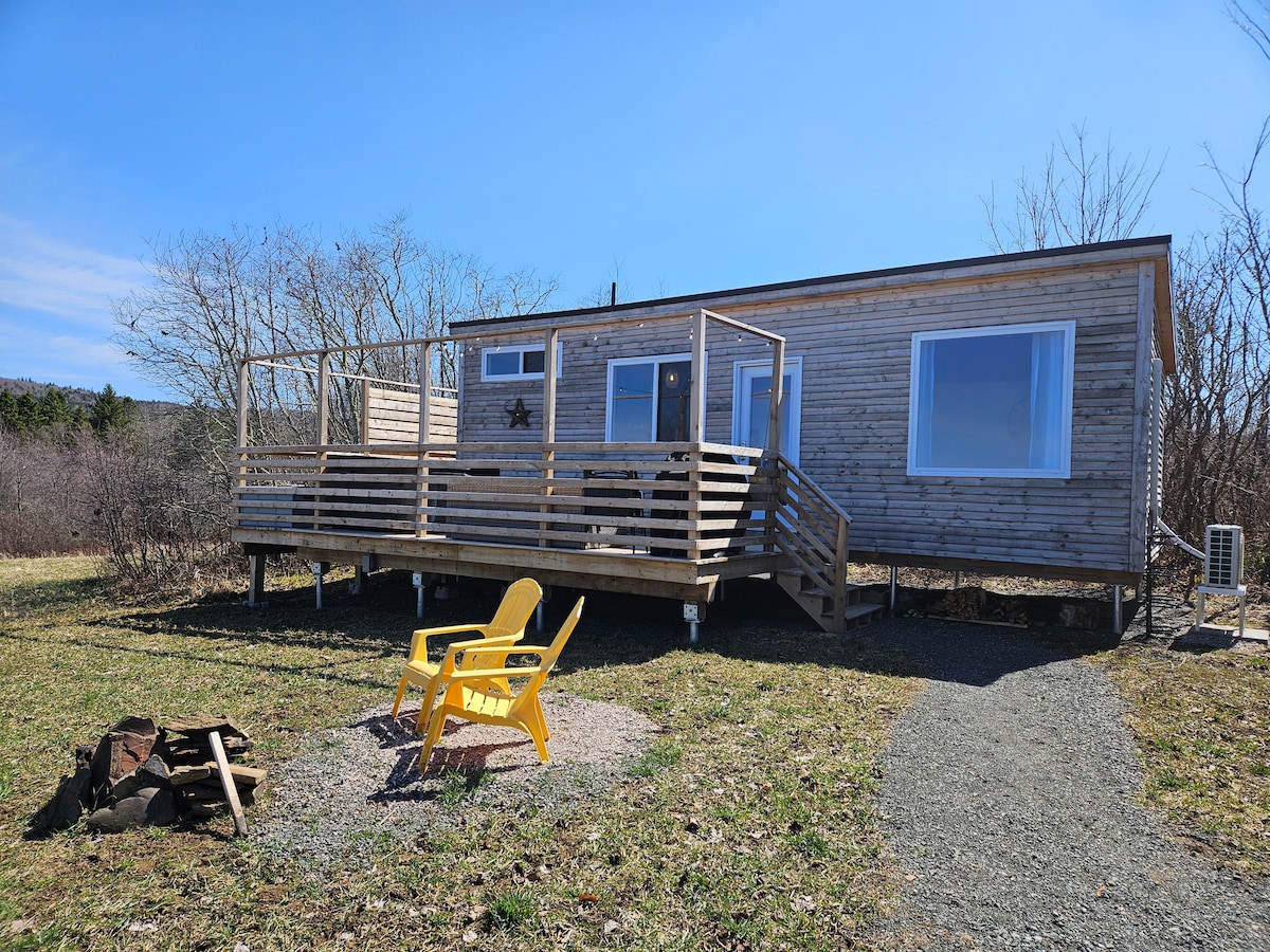 A wooden tiny home is shown featuring a spacious deck with lounge chairs. A fire pit area is visible on the ground nearby, surrounded by gravel. The building is set against a backdrop of open grass and sparse trees, with blue skies overhead.