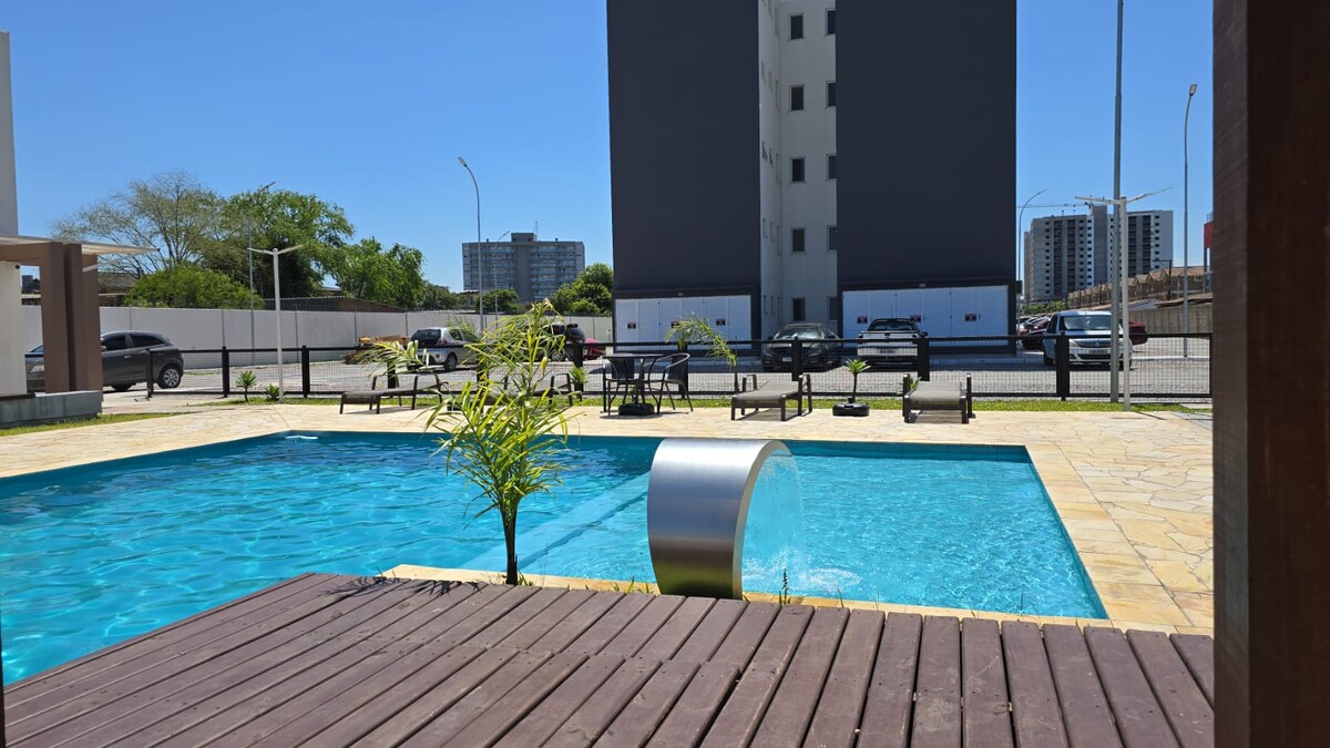 A clear blue pool is bordered by a wooden deck, reflecting sunlight under a cloudless sky. Lounge chairs are arranged around the pool, and greenery adds a natural touch to the setting. A modern building stands in the background, providing context to the tranquil atmosphere.