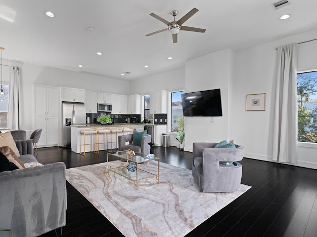 An inviting living area features a modern design with large windows allowing natural light to fill the space. Soft gray seating is arranged around a glass coffee table, with a patterned area rug underneath. A sleek television hangs on the wall, and the kitchen is visible in the background.