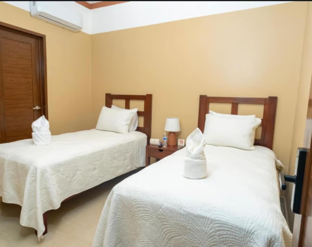 Two individual beds are positioned in a softly lit room, each with a light-coloured quilt covering. A small bedside table is situated between the beds, featuring a lamp. Warm beige walls create a calming background, complemented by a wooden door and an air conditioning unit.