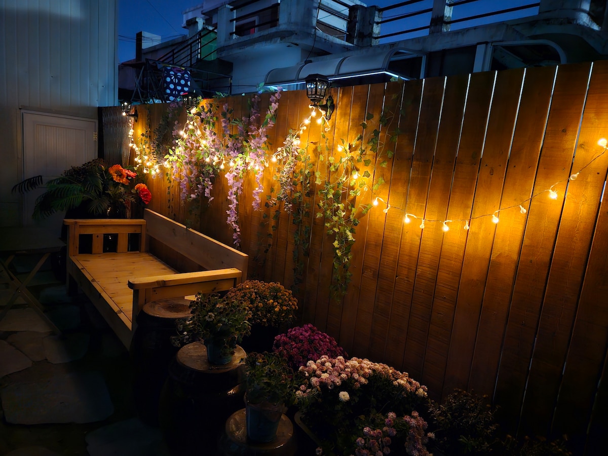 A serene outdoor space is illuminated by warm string lights, creating a relaxed ambiance. Wooden seating is complemented by vibrant flowers and greenery along the wall. The subtle glow enhances the inviting nature of the area, perfect for evening gatherings.