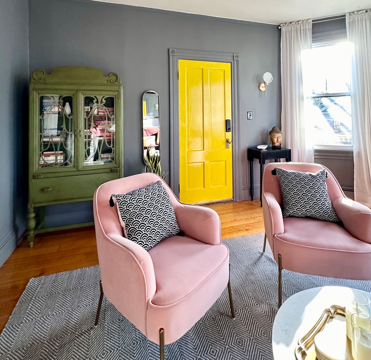 The living space is highlighted by two pink chairs positioned across from a round marble table. A green cabinet contrasts with the bold yellow door, while sheer white drapes frame a window, allowing natural light to fill the room.