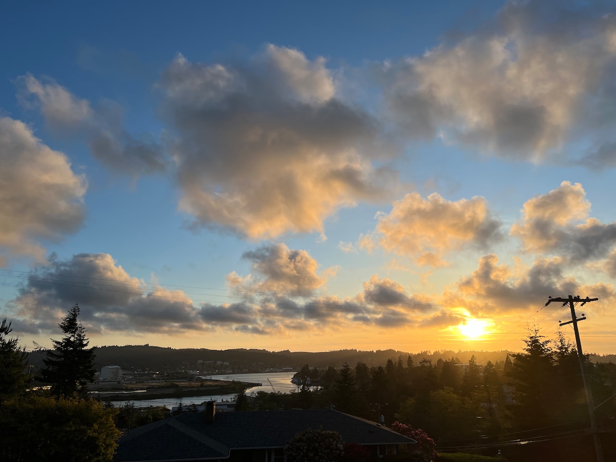 A serene sunset is displayed over the river with soft clouds painted in hues of orange and yellow. Dense trees line the riverbank, while the silhouette of the landscape creates a peaceful backdrop against the vibrant sky.