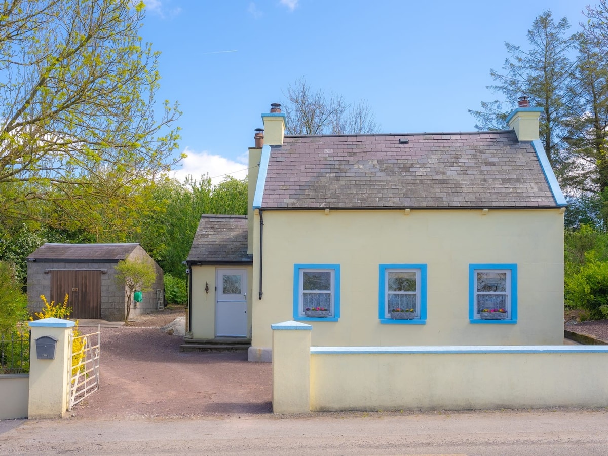 A charming cottage with a classic exterior is framed by lush greenery. The light blue trim and front door complement the yellow walls. Two front windows provide glimpses of the interior and are adorned with delicate curtains. A simple pathway leads from the gate to the entrance.