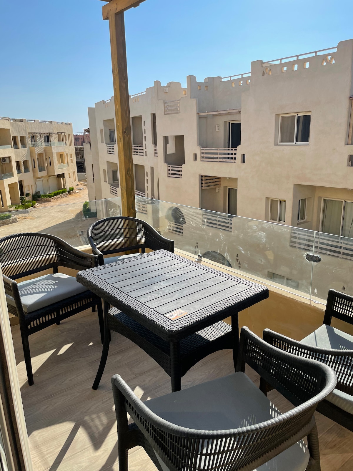 An outdoor balcony features a dark wooden table surrounded by four black wicker chairs with light cushions. The setting is bathed in sunlight, highlighting the balcony’s glass railing and offering a view of nearby residential buildings.