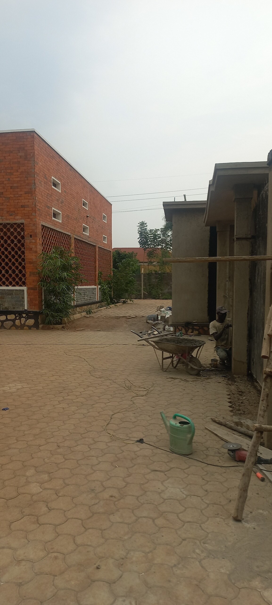 A courtyard area is visible, featuring a combination of brick and concrete structures. Plants are placed along the pathway, which is lined with patterned pavers. Tools and materials are scattered, indicating ongoing maintenance or construction.