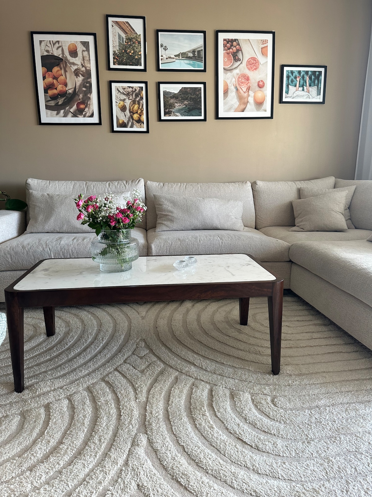 A cozy living room features a light-colored sectional sofa adorned with soft cushions. A marble-topped coffee table sits in the center, displaying a vase of fresh flowers. Framed images hang on the wall above, adding a touch of personality to the space.
