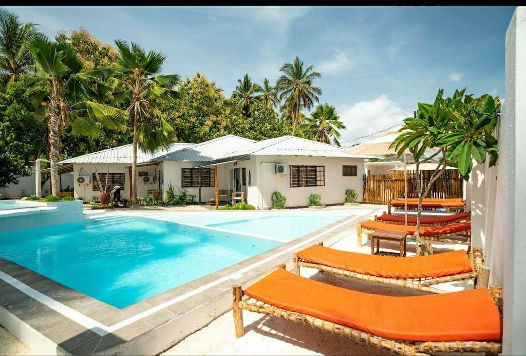 A tranquil pool area is surrounded by palm trees and tropical vegetation. Several lounge chairs with orange cushions are positioned along the poolside, inviting relaxation. The white buildings in the background feature large windows, blending seamlessly with the serene landscape.