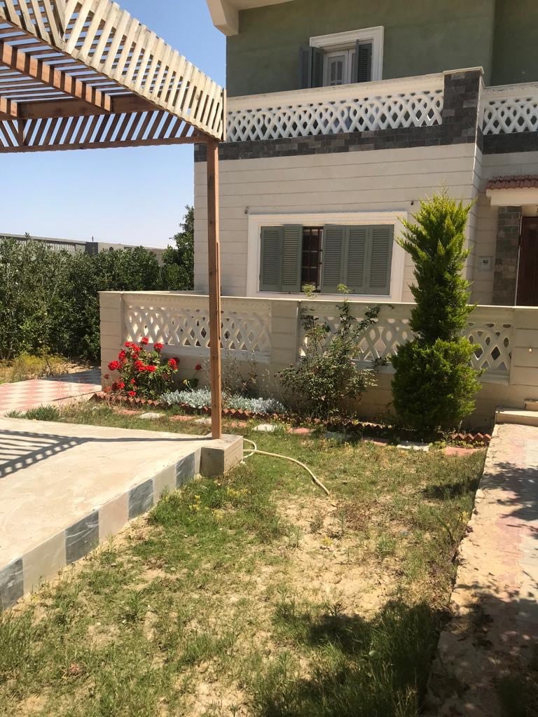 A charming outdoor space features a well-maintained garden with blooming flowers and neatly trimmed shrubs. A shaded pergola stands over a stone patio, providing a relaxing area for outdoor seating. The house reflects a light-colored exterior with a mix of traditional and modern architectural elements.