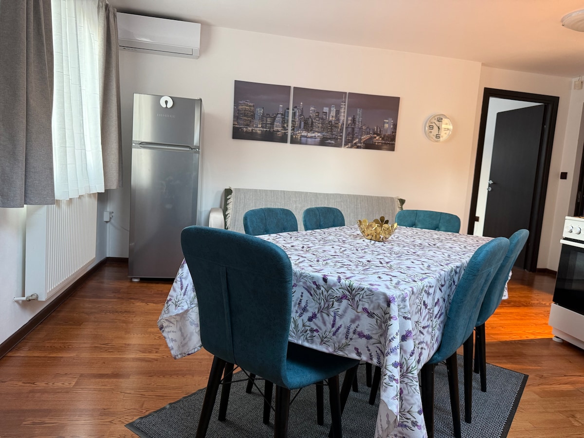 A dining area features a large table surrounded by blue upholstered chairs. A floral tablecloth covers the table. A refrigerator is positioned nearby, and artwork depicting a city skyline adorns the wall. Natural light enters through a window covered with light curtains.