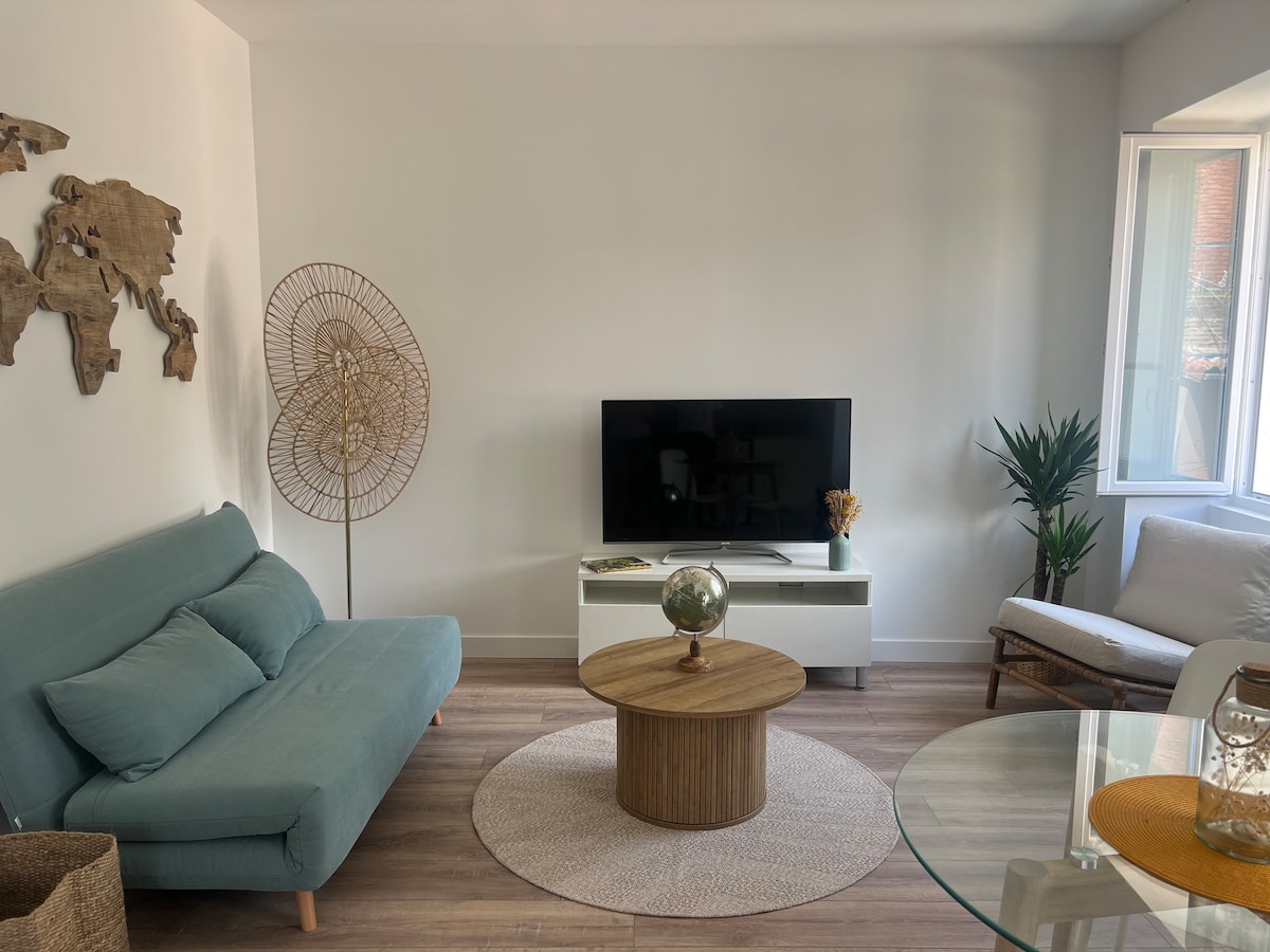 A living area is presented with a light blue sofa bed and a white television stand. A round, natural wood coffee table is centrally located on a circular rug. A potted plant and a woven floor lamp add texture, while light streams in from the windows.
